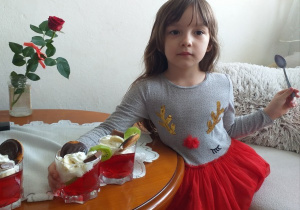w domu, dziewczynka stoi przy stole, trzyma jeden z trzech deserów, w który widać czerwoną galaretkę, bitą śmietanę, ciastko delicję i plasterek kiwi, na serwetce stoi szklany słoik z czerwoną różą